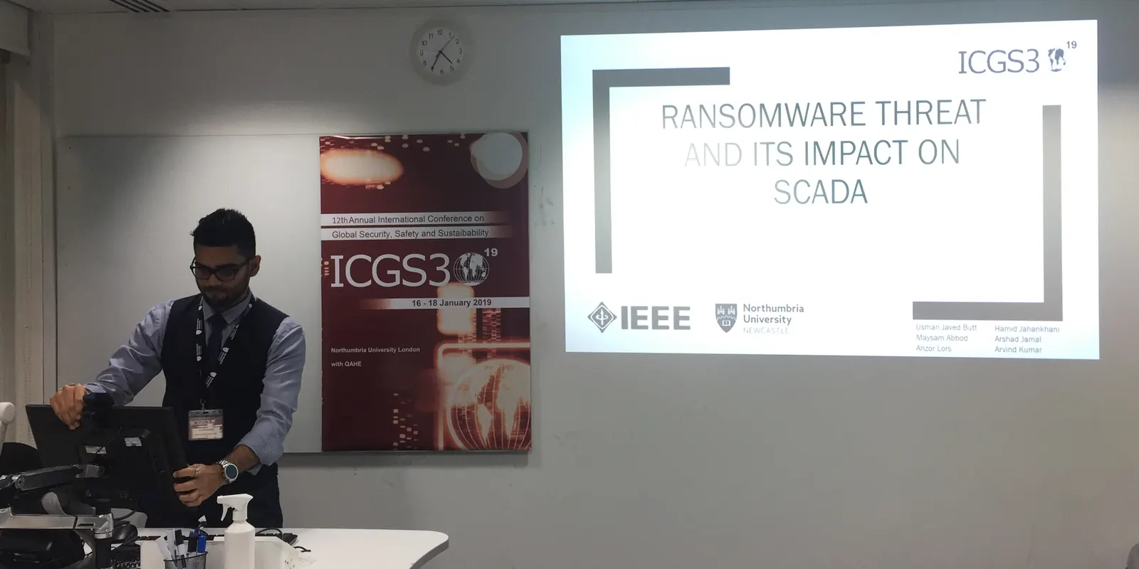 Arvind at the lectern at the IEEE ICGS3 conference, next to the conference poster, with the title slide of the SCADA paper on the screen